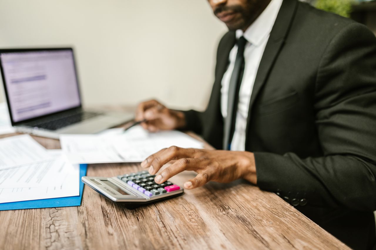 about-01 A businessman in a black suit using a calculator at a desk with financial documents and a laptop.